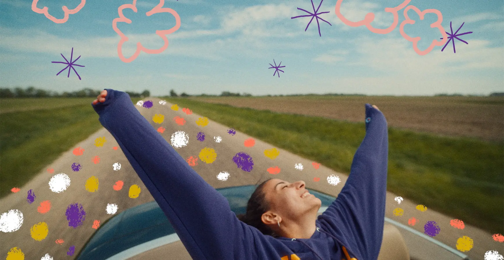 A girl rides with the back of a convertible car with her arms and head thrown back with the wind. The car is on a country road with field on both sides. Animated stars, clouds, and polka dots fill the landscape.